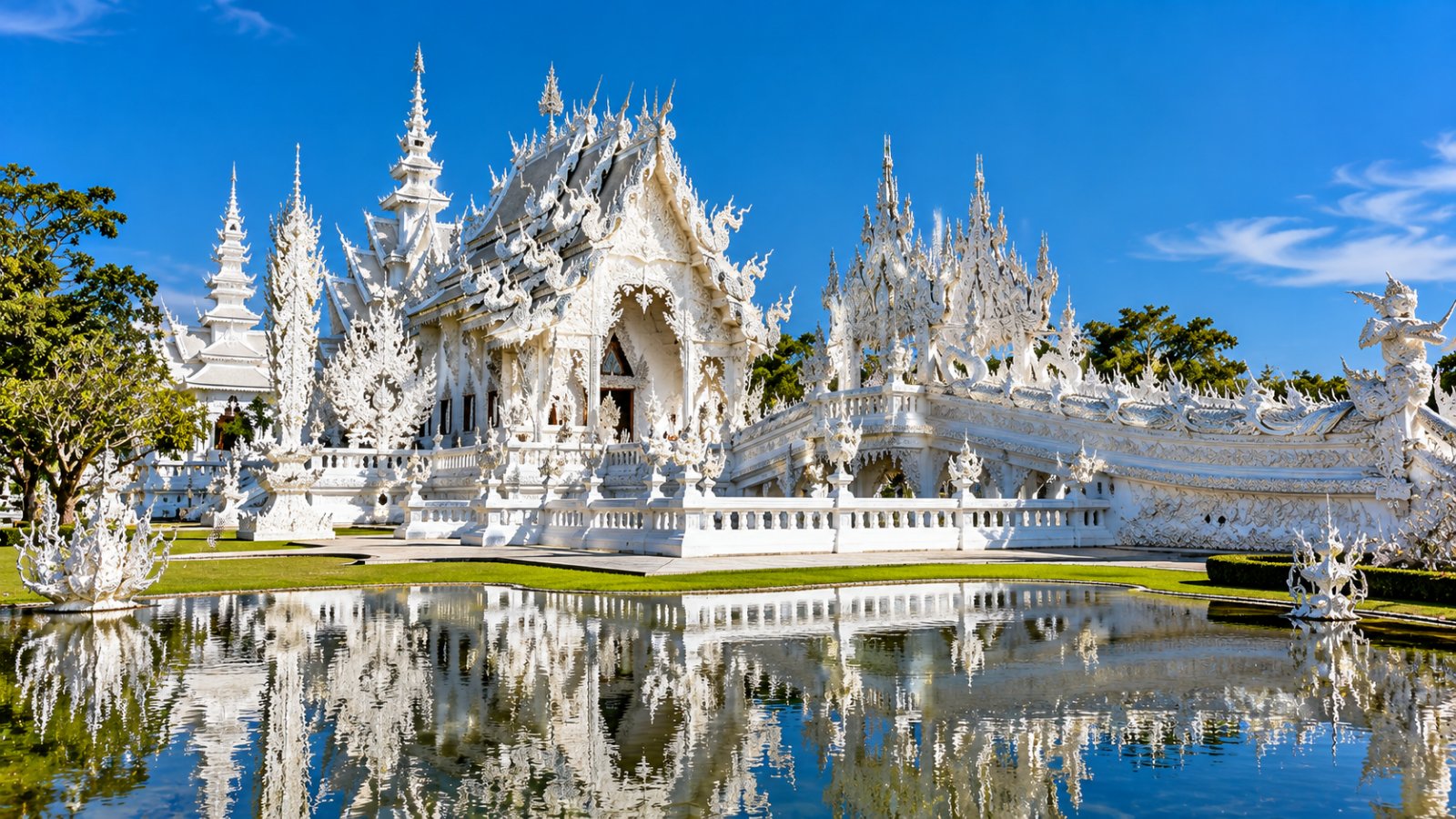 Il Tempio Bianco (Wat Rong Khun) di Chiang Rai, tutto bianco e decorato, riflesso nello stagno sotto il cielo azzurro