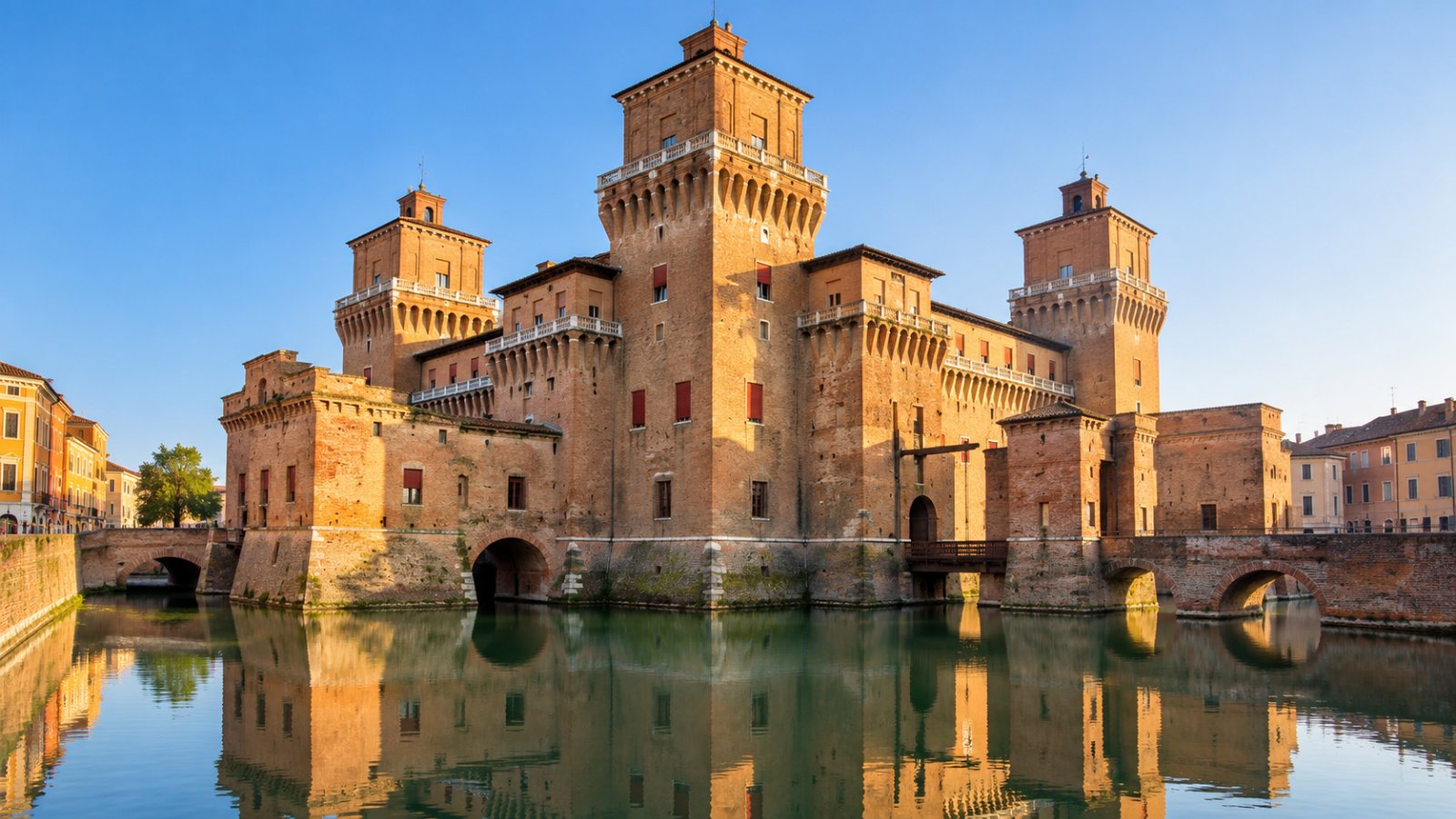 Il Castello Estense di Ferrara in mattoni rossi con le torri merlate e il fossato d'acqua in primo piano, sotto un cielo limpido di primo mattino
