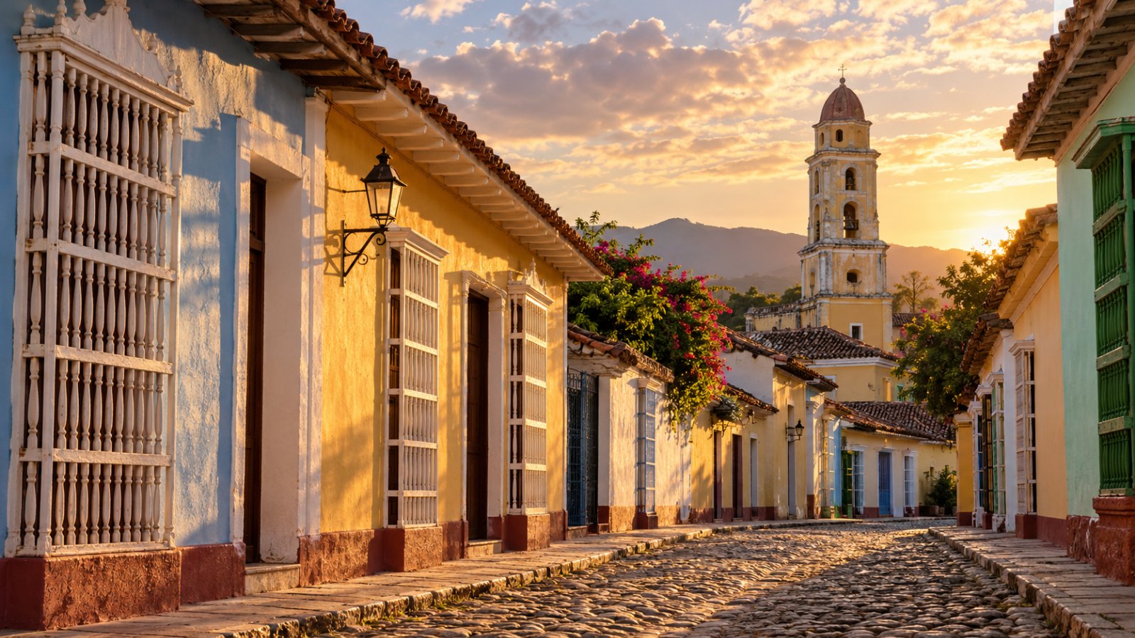 Una strada acciottolata del centro coloniale di Trinidad con case basse color pastello, grate di legno alle finestre e il campanile del convento di San Francesco sullo sfondo, al tramonto