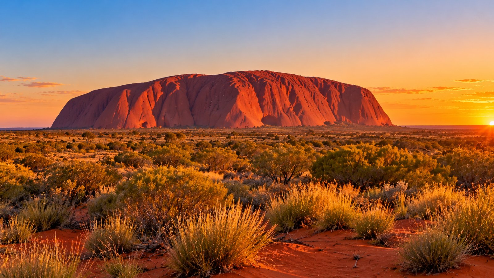 Il monolite di Uluru acceso di rosso dalla luce del tramonto, visto da lontano oltre una piana di cespugli di spinifex e dune di sabbia rossa